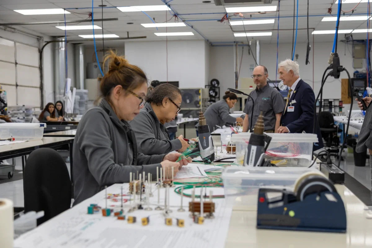 Employees assemble wire harnesses at long benches while Congressman Roger Williams tours the production floor.